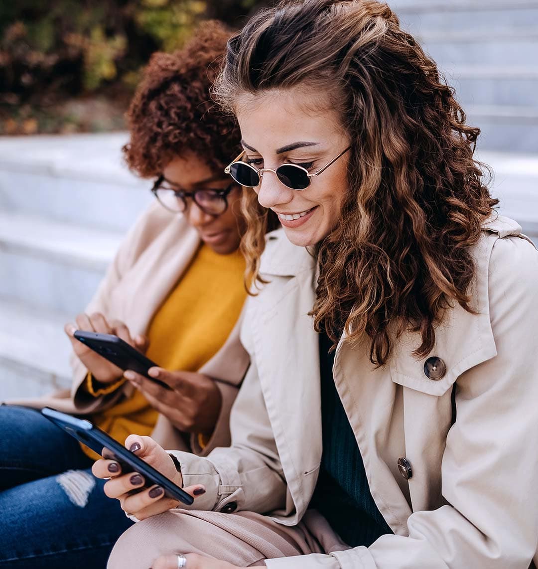 Auf dem Bild sind zwei Frauen zu sehen, die nebeneinander sitzen. Beide befinden sich an ihrem eigenen Handy und sind darauf fokussiert. Die Frau im Vordergrund lächelt.