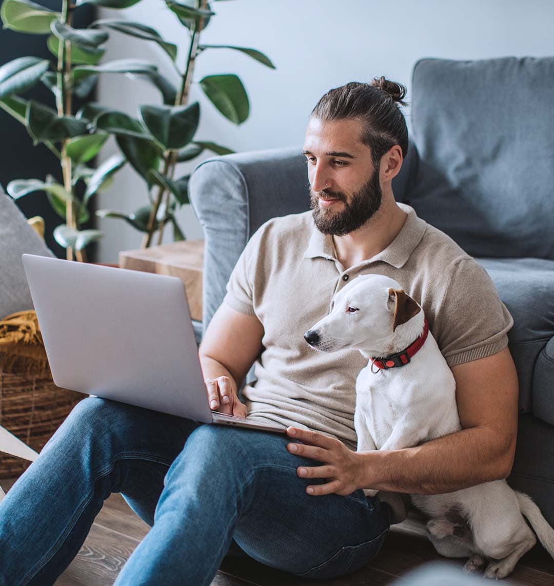 Ein Mann sitzt auf dem Boden und lehnt sich an ein Sofa an. In einem seiner Arme ist ein Hund. Beide schauen gemeinsam auf den Laptop der sich auf den Beinen des Mannes befindet.