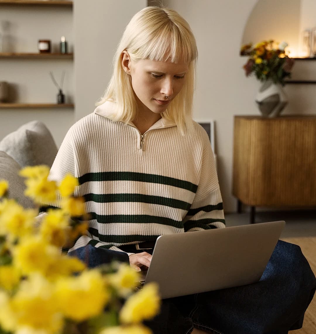 Eine junge Frau sitzt in ihrem Wohnzimmer am Laptop. Neben ihr stehen gelbe Blumen, die Atmosphäre ist ruhig.