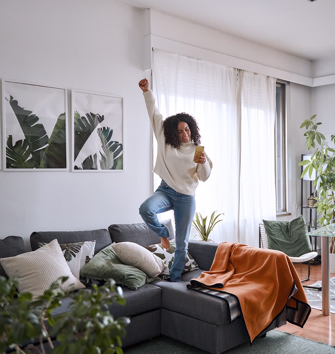 Eine junge Frau steht auf einem Bein auf einem Sofa und streckt glücklich eine Faust in die Luft. In der anderen Hand hält sie ein Smartphone, in das sie lachend schaut.