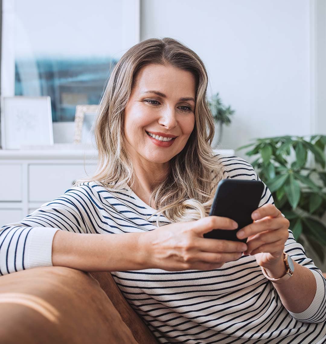 Eine Frau im Mittelpunkt des Bildes sitzt auf einem Sofa. Sie hält ein Handy in beiden Händen und schaut auf dieses. Sie lächelt dabei.