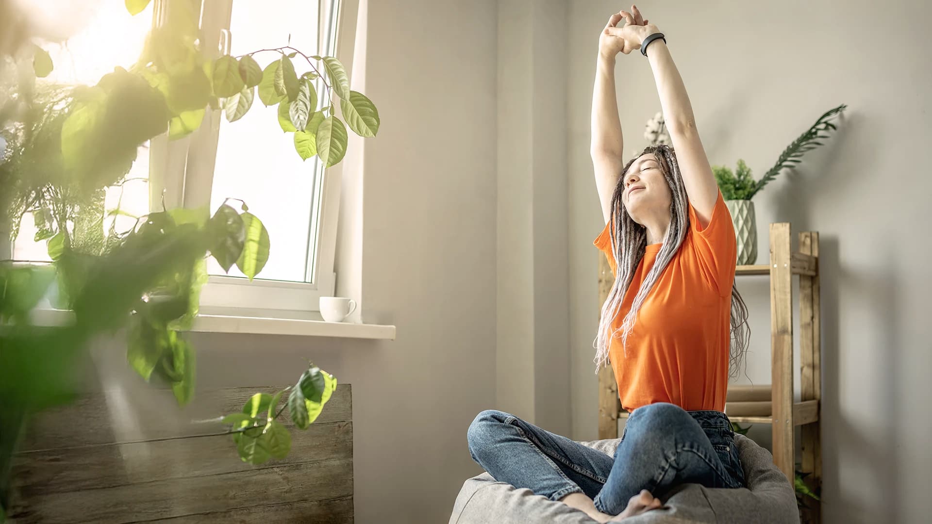 Eine junge Frau in einem orangenen Tshirt streckt sich glücklich vor einem Fenster, durch das Sonne strahlt.