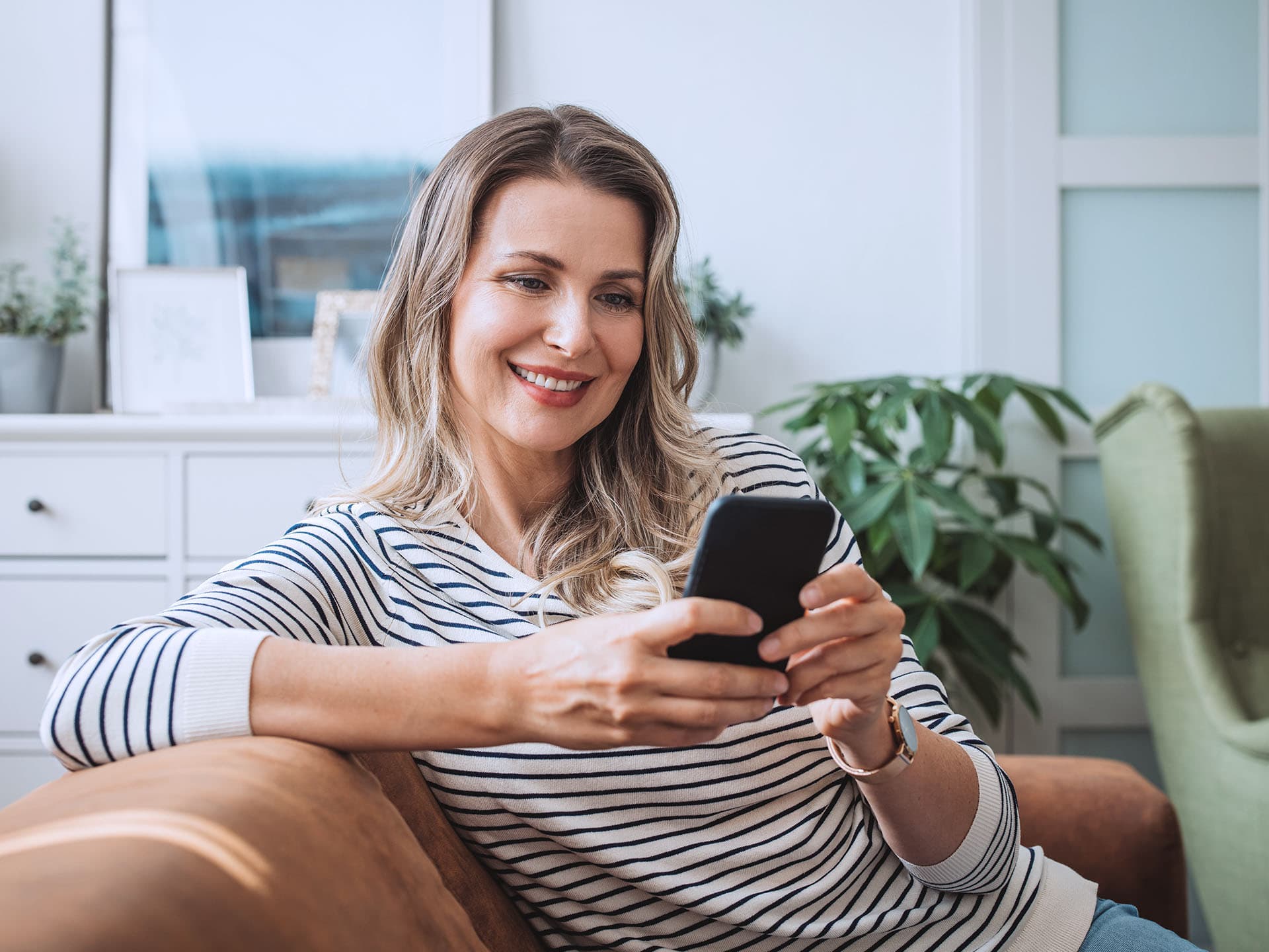 Eine Frau im Mittelpunkt des Bildes sitzt auf einem Sofa. Sie hält ein Handy in beiden Händen und schaut auf dieses. Sie lächelt dabei.