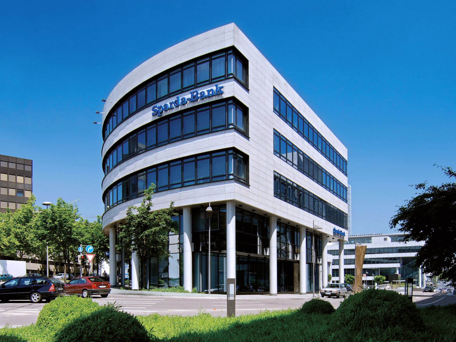 Bürogebäude der Sparda-Bank BW in Stuttgart mit großer Glasfassade, geschwungener Architektur und blauem Logo, umgeben von Bäumen und fahrenden Autos.