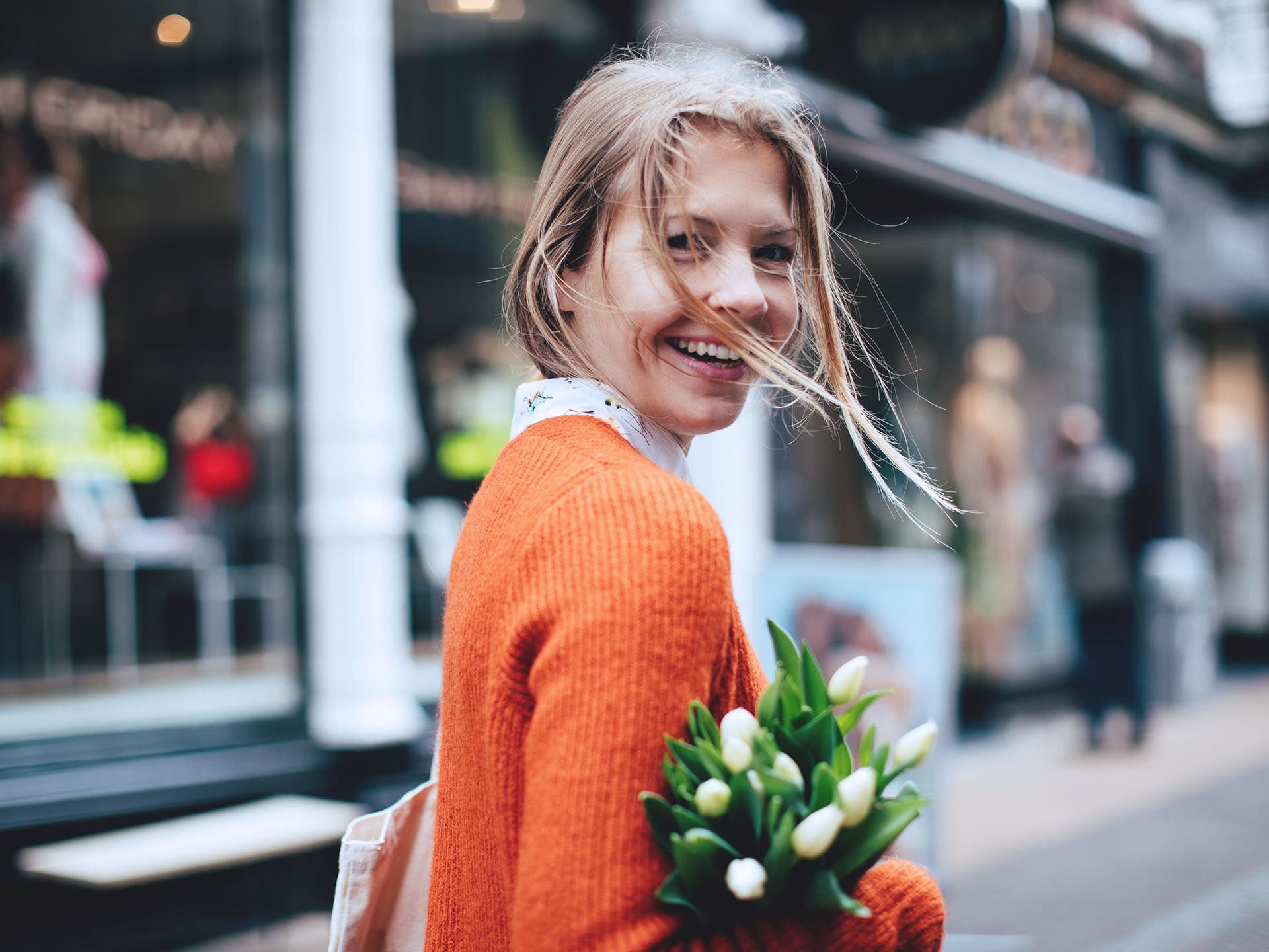 Eine Frau läuft durch eine Straße, weg von der Kamera während sie sich in die Kamera dreht und lächelt. In einem ihrer Arme befinden sich Blumen.