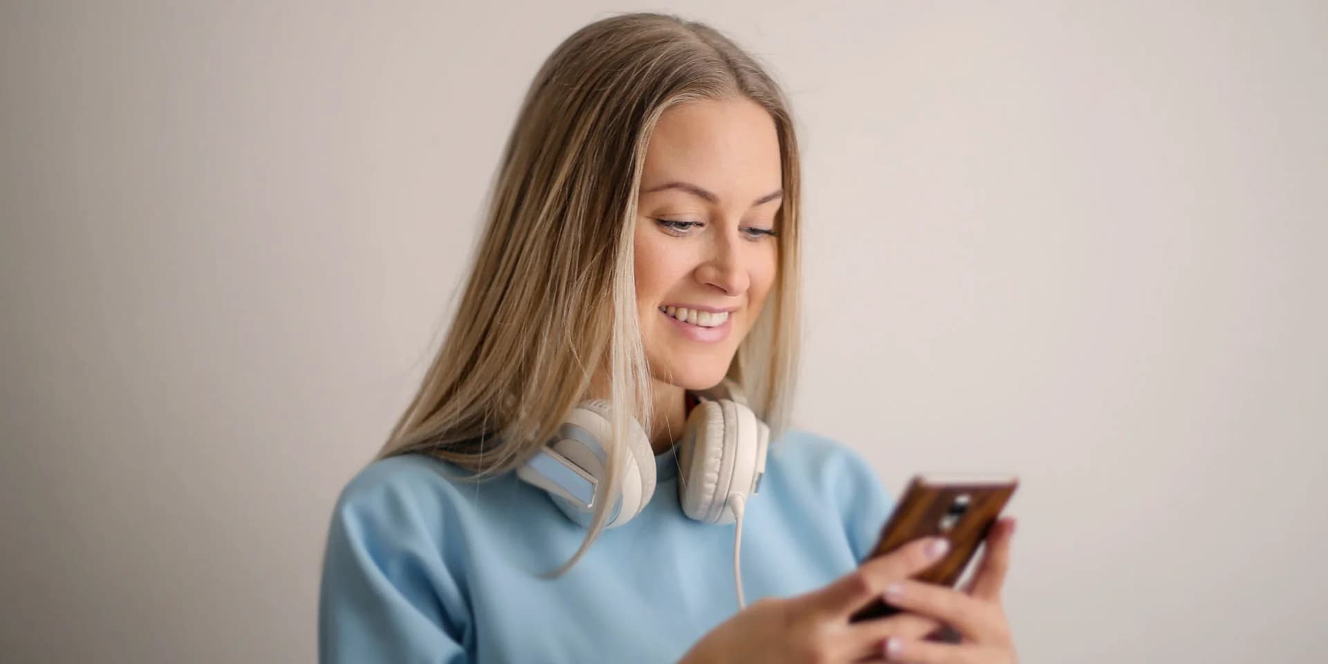 Eine junge Frau blick lächelnd auf ein Handy, welches sie vor sich hält. Um ihren Hals hängen große Kopfhörer.