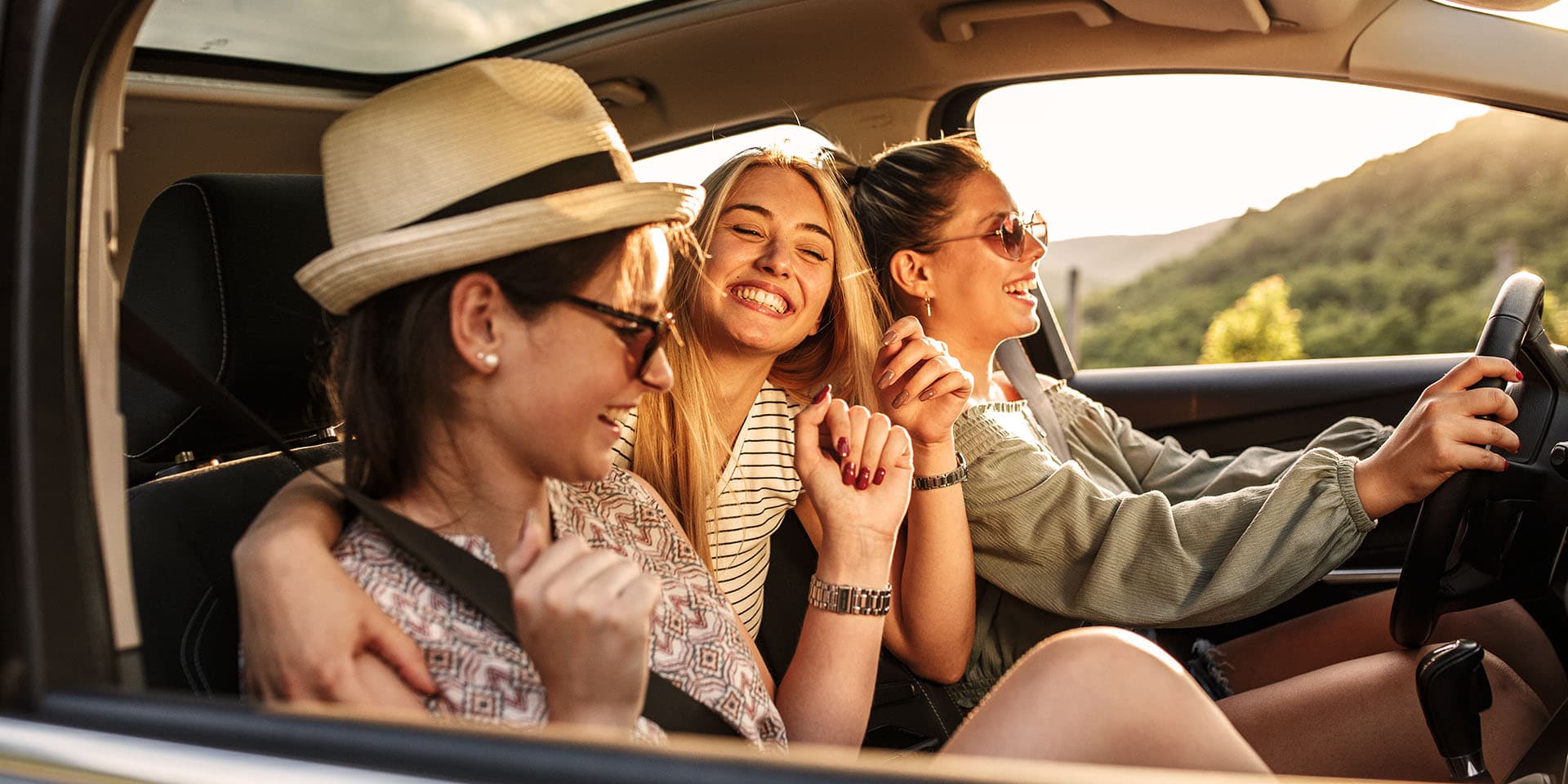 Drei Frauen sitzen in einem Auto und lachen. Aus einem der Autofenster sieht man Bäume und die Sonne, wie sie ins Auto scheint.